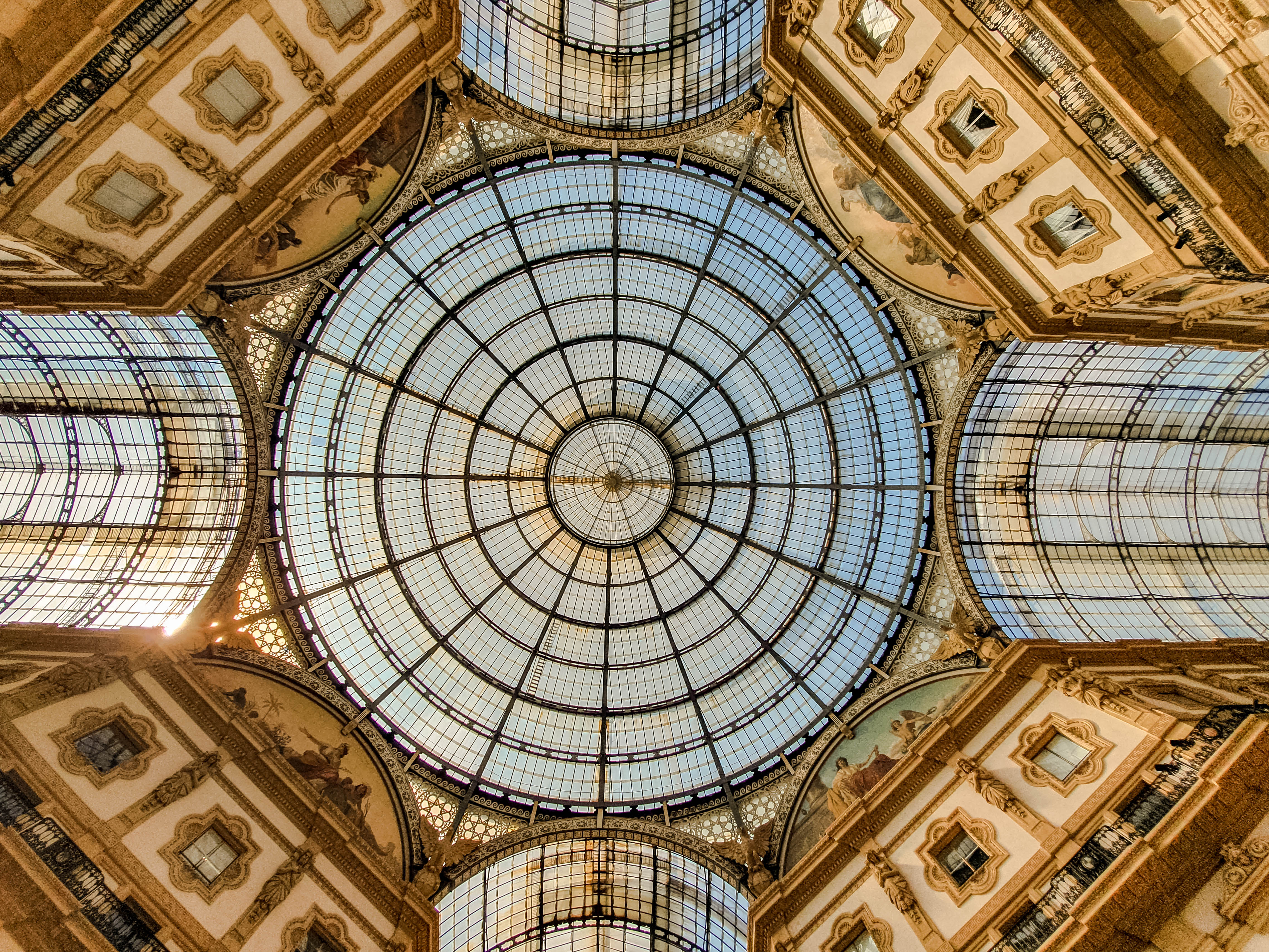 An upwards shot of the beautiful, ornate glass domed ceiling in the Galleria Vittorio Emmanuele. A clear blue sky can be seen through the glass, and the sun is peaking through the centre, left side of the dome.