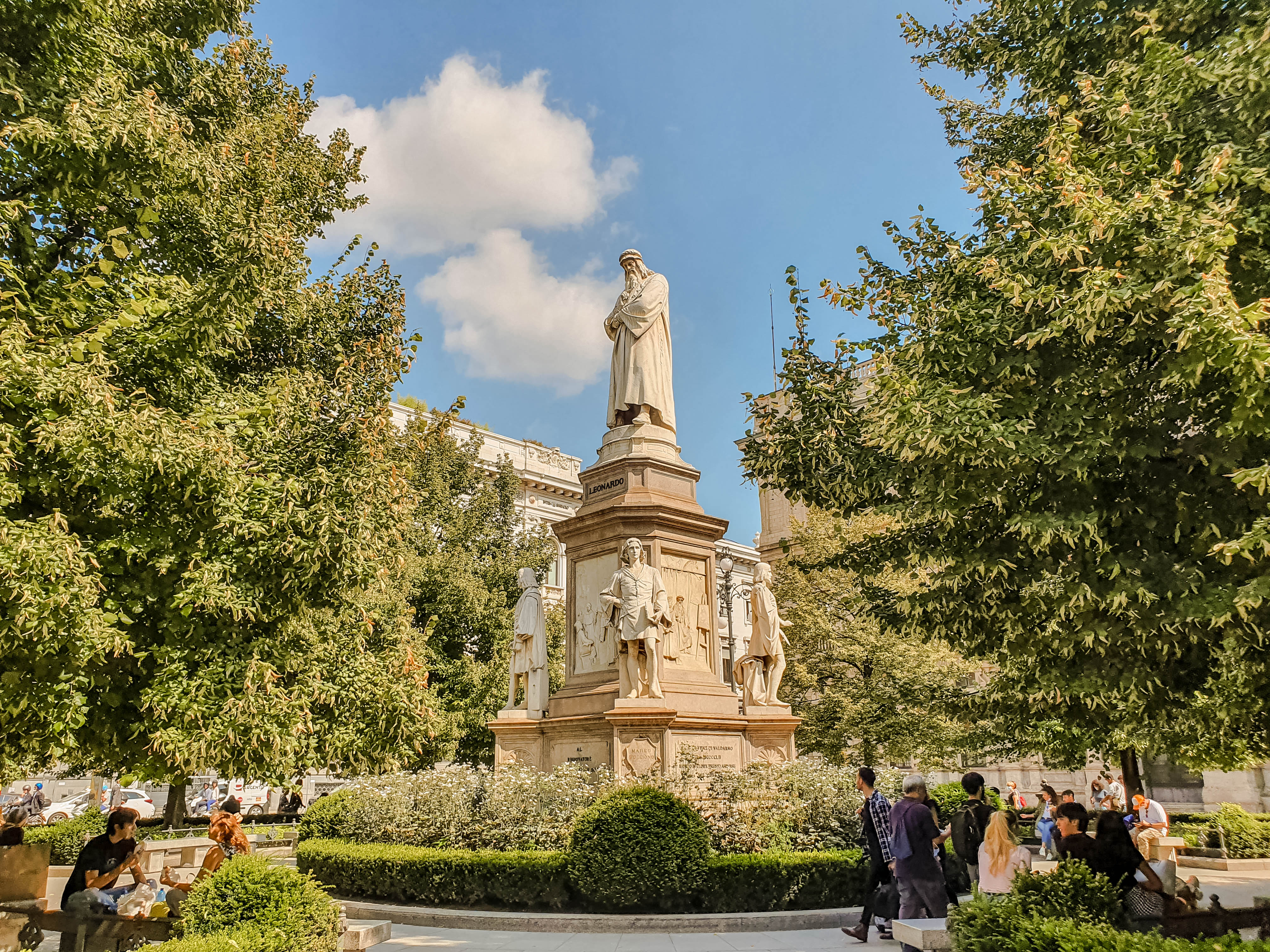 A statue of Leonardo da Vinci stands tall in a park. Greenery surrounds the statue, and people can be seen enjoying the park and sunny day.