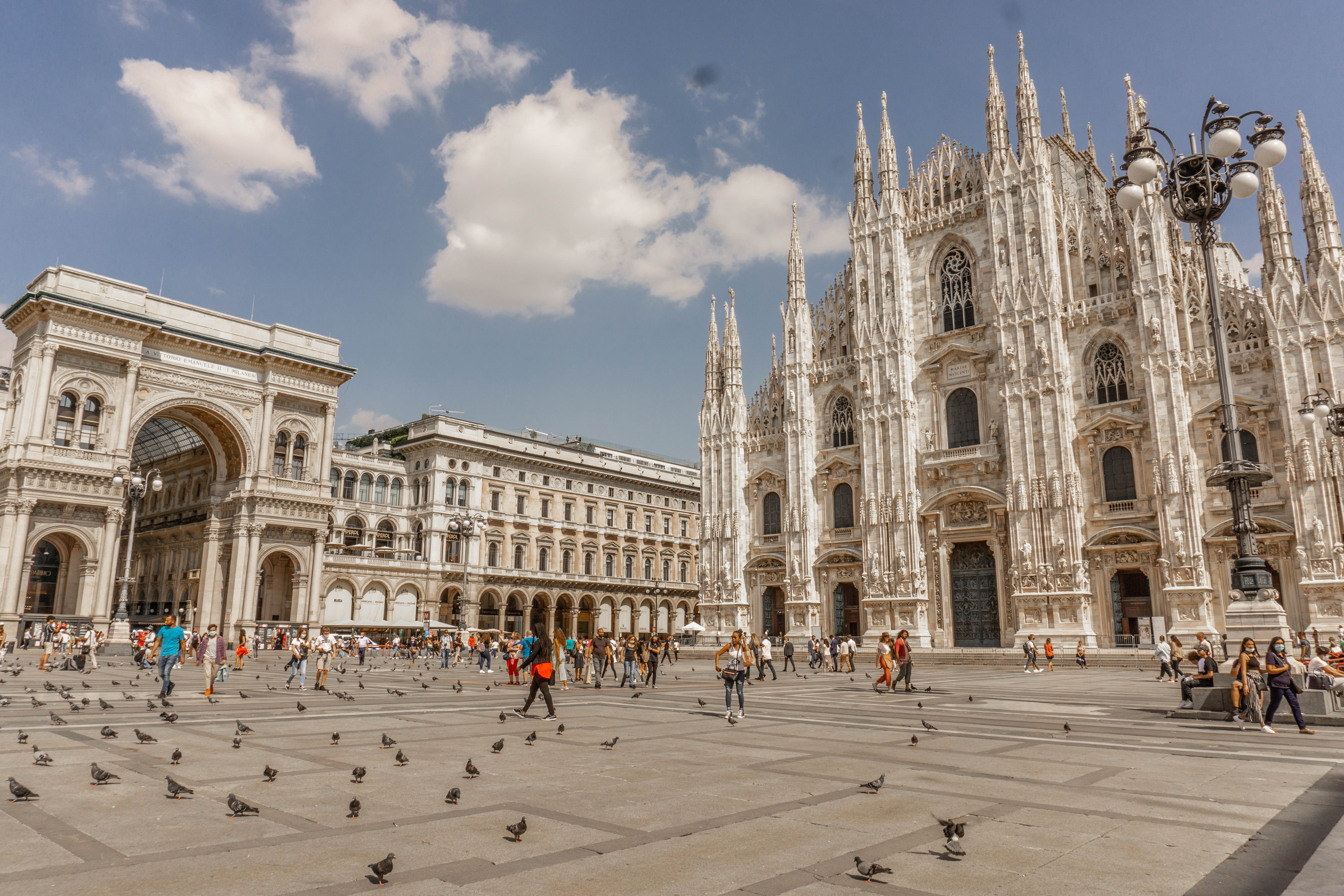 The Piazza del Duomo has a few people and birds scattered throughout the area on a sunny day. The sky is blue with clouds. The Duomo di Milano stands grandly in the right side of the shot.